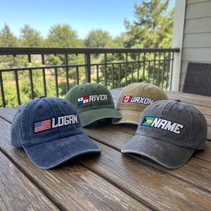 May include: Four baseball caps in various colors, each embroidered with a name and a flag. The caps are navy blue, olive green, tan, and gray. The names are "LOGAN", "RIVER", "JAXON", and "NAME". The caps are displayed on a wooden table.