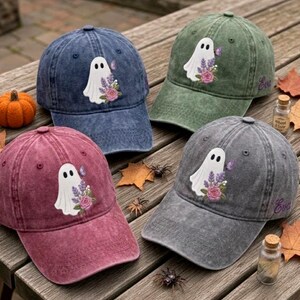 May include: Four baseball caps in blue, green, maroon, and gray, each featuring an embroidered white ghost holding a bouquet of purple flowers and a small butterfly. The green and gray caps have the name Eva embroidered on the side in purple.