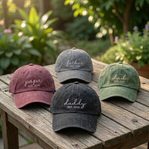 May include: Four baseball caps in various colors, each with a different word embroidered on the front: "papa," "father," "daddy," and "dad." Each hat also has "EST. 2026" embroidered below the word. The hats are arranged on a weathered wooden surface.