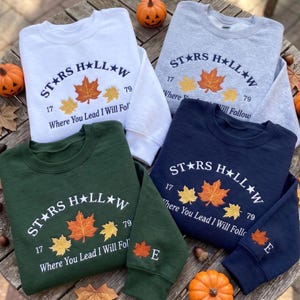 May include: Four crewneck sweatshirts in white, light gray, dark green, and navy blue. Each features the text Stars Hollow, 1779, and Where You Lead I Will Follow, with three embroidered maple leaves. The green and navy sleeves show an E and a leaf.