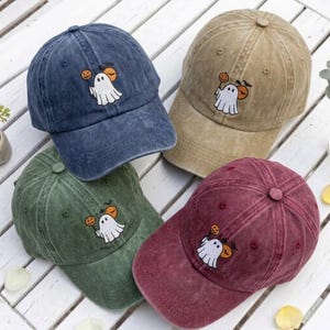 May include: Four baseball caps in blue, tan, green, and maroon, each featuring a washed-out fabric texture. Every cap displays an embroidered design of a white ghost holding a small orange pumpkin, accompanied by a black bat silhouette above.