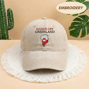 May include: A beige baseball cap with the text "HANDS OFF GREENLAND" and a Greenland map graphic in red and white. The cap is sitting on a white, fringed mat. The word "EMBROIDERY" is in a circle in the background.