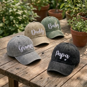 May include: Four baseball caps in various colors: gray, tan, olive green, and black. Each cap has white embroidered text: "Gramps", "Daddy", "Baba", and "Papa". The caps are displayed on a wooden surface with plants in the background.