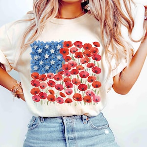 May include: A cream-coloured t-shirt with a floral American flag design. The flag's stars are white, the stripes are red poppies, and the blue field is made of blue flowers. The shirt is worn with blue denim jeans.