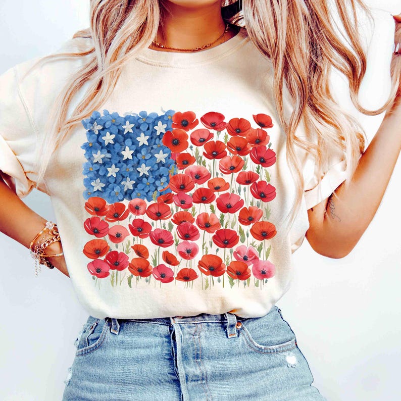May include: A cream-coloured t-shirt with a floral American flag design. The flag's stars are white, the stripes are red poppies, and the blue field is made of blue flowers. The shirt is worn with blue denim jeans.