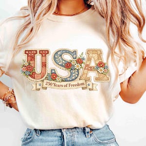May include: Cream-colored t-shirt featuring the word "USA" in a decorative, patchwork style. The letters are adorned with floral accents and a banner reading "250 Years of Freedom."