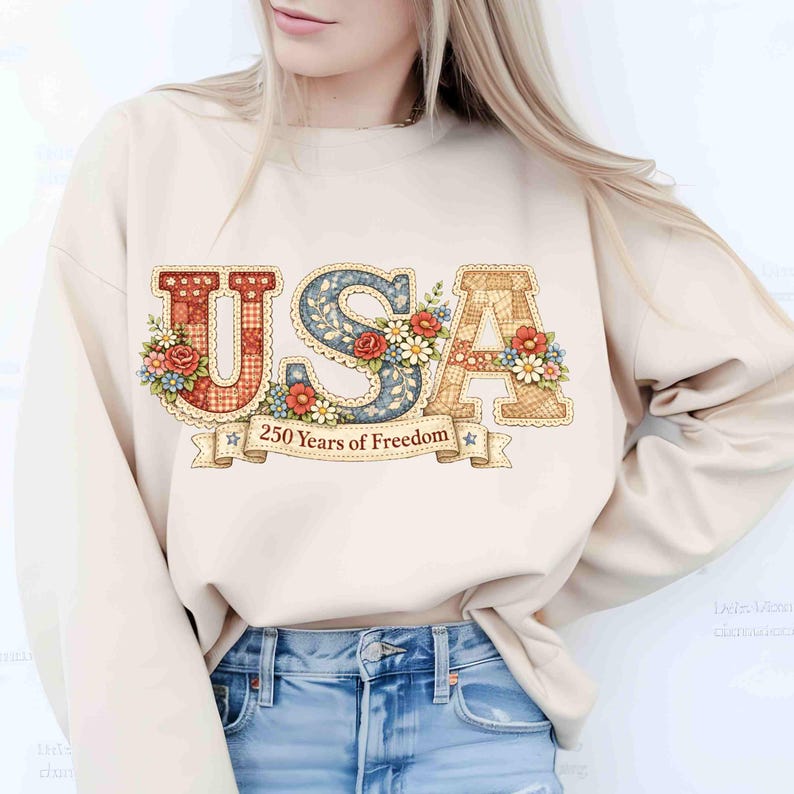May include: Cream-colored sweatshirt with a patriotic design. The word "USA" is displayed in a decorative font, adorned with floral accents. A banner below reads "250 Years of Freedom". The design features red, blue, and tan colors.