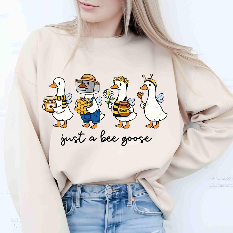 May include: Beige sweatshirt featuring a whimsical illustration of four geese dressed as bees, each holding honey-related items. The text "just a bee goose" is printed below the geese in a black script font.