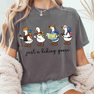 K&ouml;nnte beinhalten: Graues T-Shirt mit einer Illustration von vier Cartoon-G&auml;nsen, die zum Wandern ausger&uuml;stet sind, jede mit Rucksack und Ausr&uuml;stung. Der Text "just a hiking goose" ist darunter in Schreibschrift geschrieben.