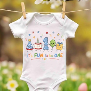 May include: A white short-sleeved baby bodysuit featuring a colorful graphic of a party hat, a birthday cake, the number one, a green balloon, and a gift box with the text it's FUN to be ONE, surrounded by small stars and confetti.