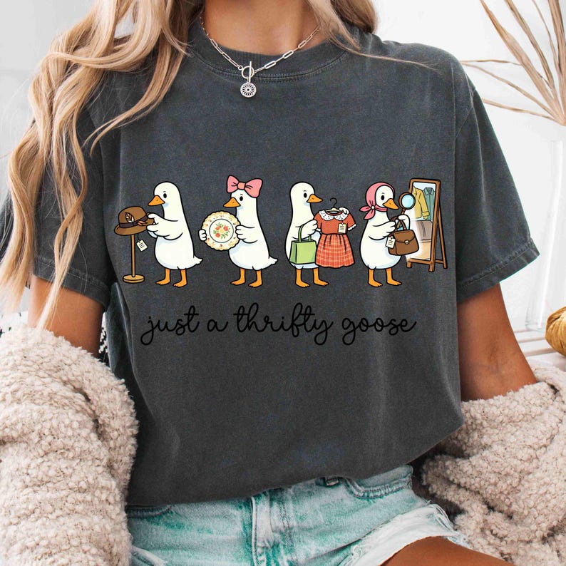 May include: Dark grey t-shirt with a cartoon design of geese, each carrying items such as a hat, plate, dress, and handbag. The cursive text below reads "just a thrifty goose".