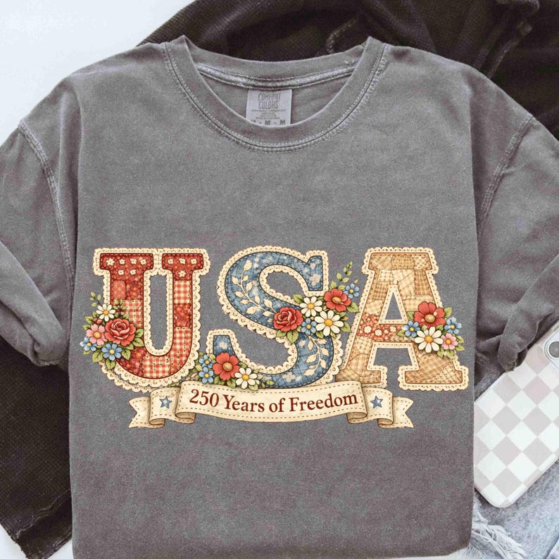 May include: Gray long-sleeve shirt with the word "USA" in a decorative font. The letters are adorned with floral patterns and surrounded by a banner that reads "250 Years of Freedom". The shirt has a vintage, washed look.