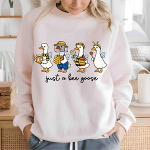 May include: A light pink sweatshirt featuring a whimsical illustration of four cartoon geese dressed as bees. Each goose holds honey-related items. The text "just a bee goose" is printed below the geese in black script.