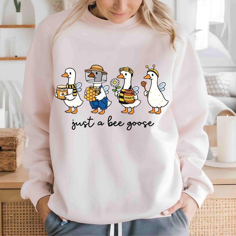 May include: A light pink sweatshirt featuring a whimsical illustration of four cartoon geese dressed as bees. Each goose holds honey-related items. The text "just a bee goose" is printed below the geese in black script.