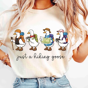 K&ouml;nnte beinhalten: Cremefarbenes T-Shirt mit einer Illustration von vier G&auml;nsen, die zum Wandern ausger&uuml;stet sind, jede mit Rucksack und Ausr&uuml;stung. Der Text "just a hiking goose" ist in kursiver Schrift unter den G&auml;nsen geschrieben. Die G&auml;nse tragen H&uuml;te und Stiefel.