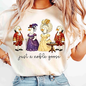 Puede incluir: Camiseta color crema con una ilustración humorística de cuatro gansos vestidos con trajes históricos. Un ganso está jugando al ajedrez. El texto "just a noble goose" está escrito debajo, añadiendo un toque de humor al diseño.
