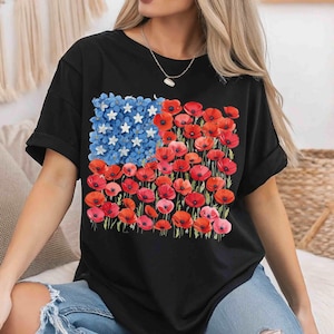 May include: Black t-shirt with a floral American flag design. The flag features blue flowers with white star accents and red poppies. The shirt has short sleeves and a relaxed fit.