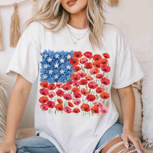 May include: White t-shirt with a floral American flag design. The flag features blue flowers and white stars in the upper left, with red poppies and pink flowers forming the stripes. A casual, patriotic tee.
