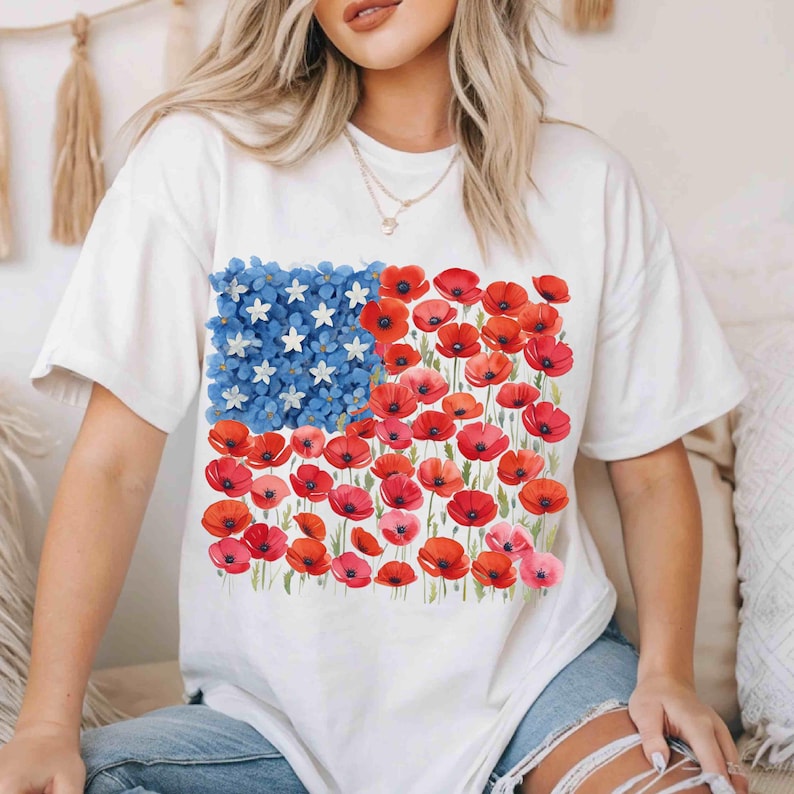 May include: White t-shirt with a floral American flag design. The flag features blue flowers and white stars in the upper left, with red poppies and pink flowers forming the stripes. A casual, patriotic tee.