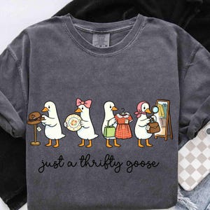 May include: Dark grey sweatshirt featuring a cartoon of geese shopping. The geese are depicted with various items: a hat, a plate, a dress, and a handbag. The text "just a thrifty goose" is written in cursive below the geese.
