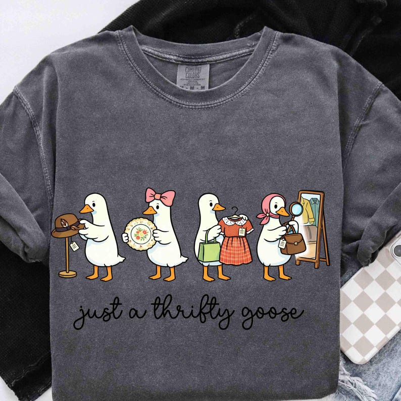 May include: Dark grey sweatshirt featuring a cartoon of geese shopping. The geese are depicted with various items: a hat, a plate, a dress, and a handbag. The text "just a thrifty goose" is written in cursive below the geese.