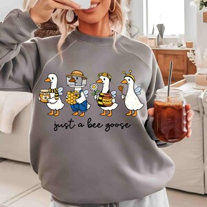 May include: Gray sweatshirt featuring a whimsical design of four geese dressed as bees, each holding honey-related items. The phrase "just a bee goose" is printed below the geese in a playful font. The sweatshirt is a comfortable, casual style.