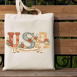May include: Cream-colored tote bag featuring the word "USA" in a decorative font, adorned with floral accents. The text "250 Years of Freedom" is displayed on a banner below. The bag has long handles.