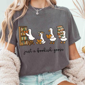 May include: Dark gray t-shirt featuring a cartoon of four geese reading books. One goose is at a bookshelf, another is sitting, and the last is carrying a stack of books. The text "just a bookish goose" is below the geese.