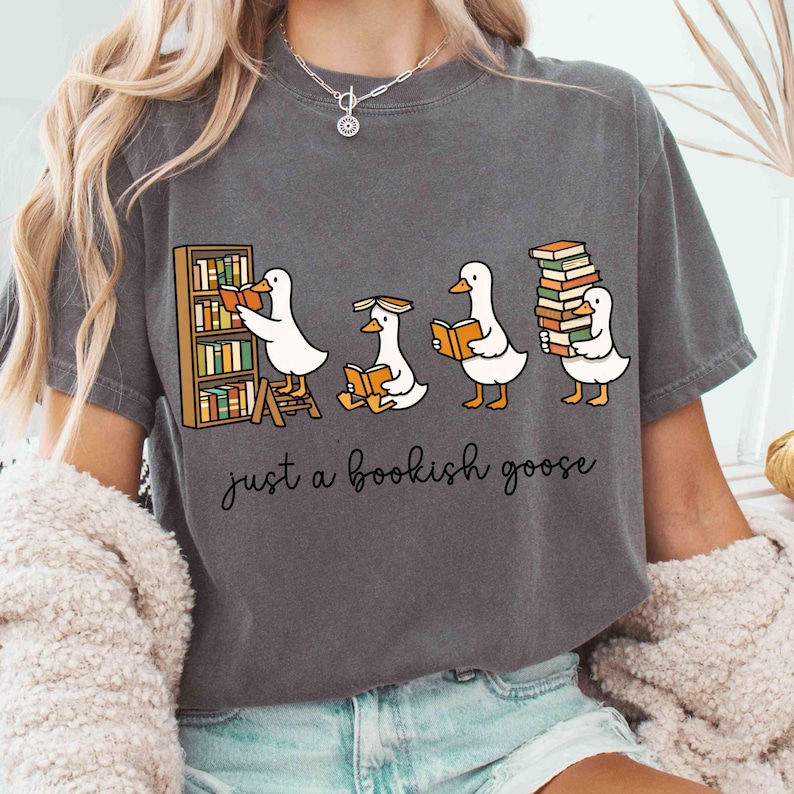 May include: Dark gray t-shirt featuring a cartoon of four geese reading books. One goose is at a bookshelf, another is sitting, and the last is carrying a stack of books. The text "just a bookish goose" is below the geese.