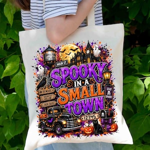 May include: A white canvas tote bag featuring a colourful Halloween graphic with a black truck, a haunted house, a water tower, pumpkins, a black cat, and the text "SPOOKY IN A SMALL TOWN" in purple and orange lettering with a "12345" sign.