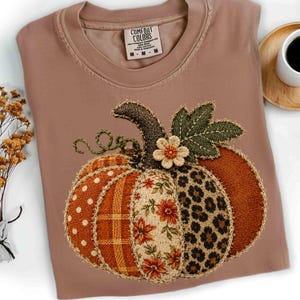 Peut inclure: Un t-shirt rose poudré avec une étiquette Comfort Colors au col, arborant un grand motif de citrouille. La citrouille est composée de divers segments texturés incluant des pois, des carreaux, des motifs floraux et un imprimé léopard, surmontée d'une fleur.