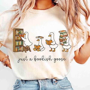 May include: Cream-colored t-shirt featuring a whimsical illustration of four cartoon geese reading books. One goose is on a ladder, another is sitting, and the last is carrying a stack of books. The text "just a bookish goose" is written below the geese.