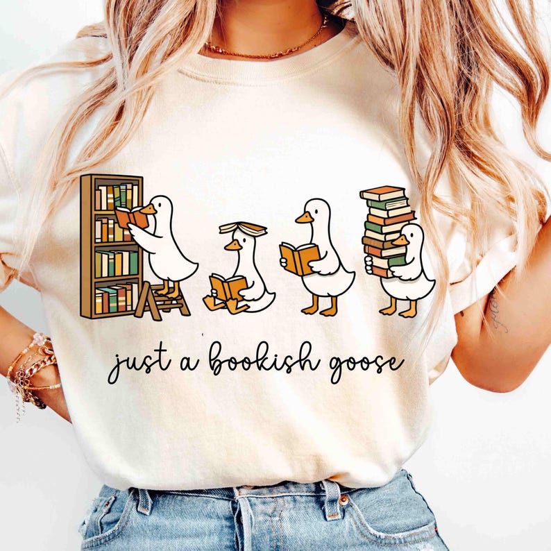May include: Cream-colored t-shirt featuring a whimsical illustration of four cartoon geese reading books. One goose is on a ladder, another is sitting, and the last is carrying a stack of books. The text "just a bookish goose" is written below the geese.