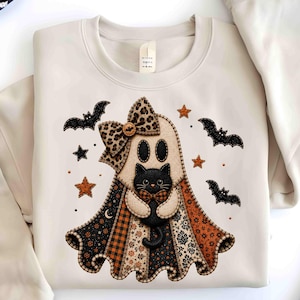 May include: A beige crewneck sweatshirt featuring a central graphic of a ghost wearing a leopard print bow and holding a black cat. The ghost is draped in a patchwork-style cape with orange, black, and white patterns, surrounded by bats and stars.