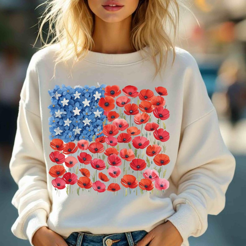 May include: Cream-coloured sweatshirt with a floral American flag design. The flag features blue forget-me-nots and white stars, with red poppies forming the stripes. The sweatshirt has long sleeves and a crew neck.