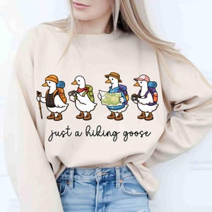 K&ouml;nnte beinhalten: Beiges Sweatshirt mit einer skurrilen Illustration von vier Comic-G&auml;nsen, die jeweils mit Wanderausr&uuml;stung ausgestattet sind. Die G&auml;nse sind mit Rucks&auml;cken, H&uuml;ten und Kameras dargestellt, mit dem Schriftzug "just a hiking goose" darunter.