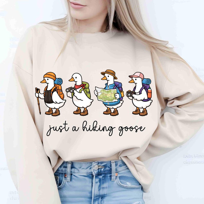 K&ouml;nnte beinhalten: Beiges Sweatshirt mit einer skurrilen Illustration von vier Comic-G&auml;nsen, die jeweils mit Wanderausr&uuml;stung ausgestattet sind. Die G&auml;nse sind mit Rucks&auml;cken, H&uuml;ten und Kameras dargestellt, mit dem Schriftzug "just a hiking goose" darunter.