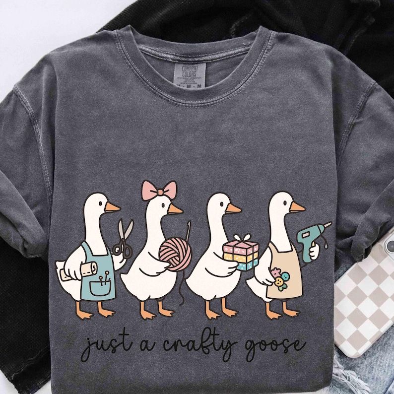 May include: Dark gray long-sleeve shirt with a whimsical illustration of four geese, each holding craft supplies. One goose has scissors, another yarn, a third a gift, and the last a glue gun. The text reads "just a crafty goose."