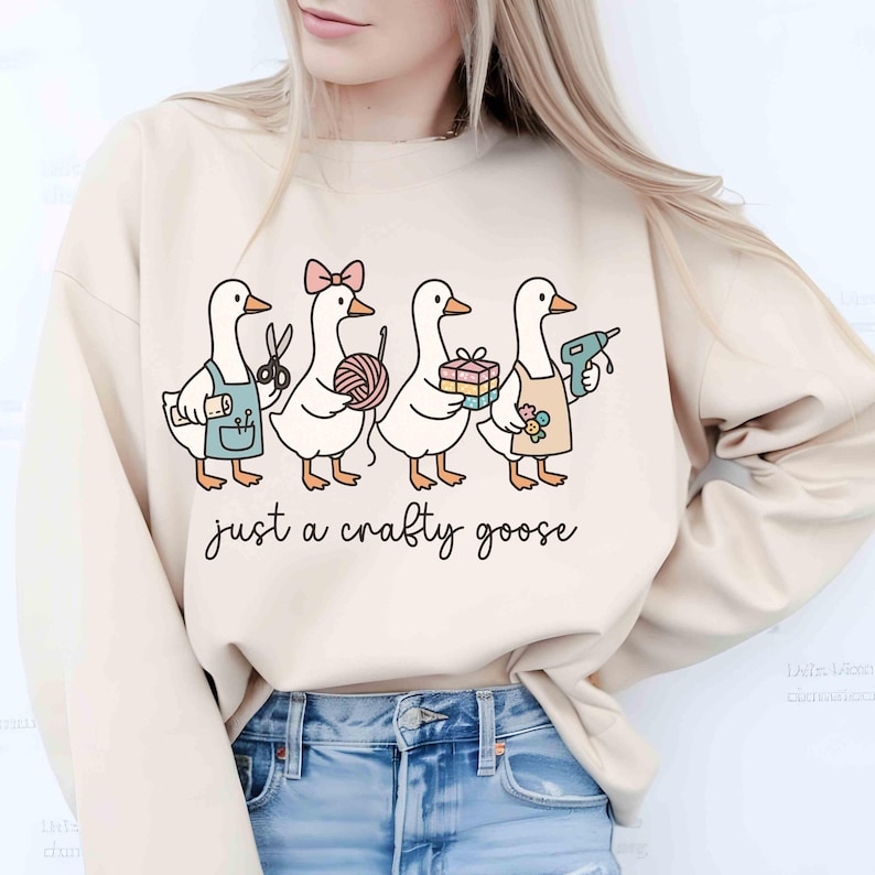 May include: Beige sweatshirt featuring a whimsical illustration of four geese, each holding craft supplies like scissors, yarn, and a glue gun. The text "just a crafty goose" is written below the geese in a handwritten style font.