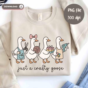 May include: Beige sweatshirt featuring a whimsical illustration of four geese, each holding craft supplies like scissors, yarn, and a glue gun. The text "just a crafty goose" is written below the geese. The image also includes the text "PNG file 300 dpi".