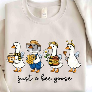 May include: Beige sweatshirt featuring a whimsical illustration of four geese dressed as bees. Each goose holds honey-related items, such as a jar of honey, honeycomb, and a honey dipper. The text "just a bee goose" is written in a cursive font below the illustration.
