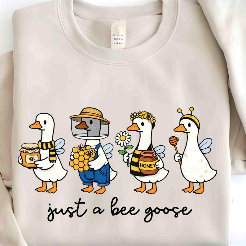 May include: Beige sweatshirt featuring a whimsical illustration of four geese dressed as bees. Each goose holds honey-related items, such as a jar of honey, honeycomb, and a honey dipper. The text "just a bee goose" is written in a cursive font below the illustration.