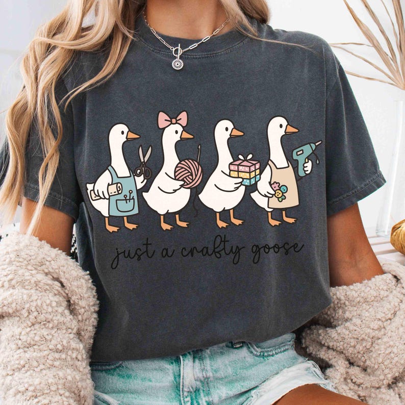 May include: Dark gray t-shirt featuring a whimsical illustration of four geese, each holding craft supplies like scissors, yarn, and a glue gun. The text "just a crafty goose" is printed below the geese.