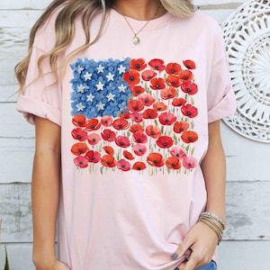 May include: A pale pink t-shirt with a floral American flag design. The flag's stars are white flowers, and the stripes are red poppies. The shirt has rolled-up sleeves and a crew neck.