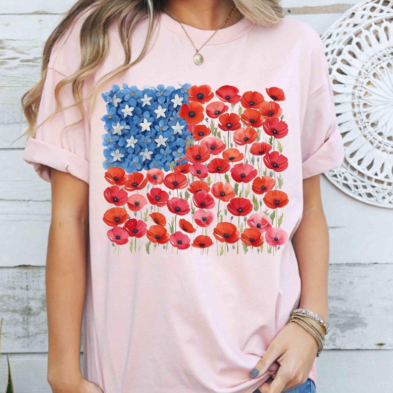 May include: A pale pink t-shirt with a floral American flag design. The flag's stars are white flowers, and the stripes are red poppies. The shirt has rolled-up sleeves and a crew neck.
