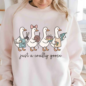 May include: A light pink sweatshirt featuring a whimsical illustration of four geese, each holding craft supplies. One goose has scissors, another yarn, a third a gift, and the last a glue gun. The text "just a crafty goose" is written below the geese.