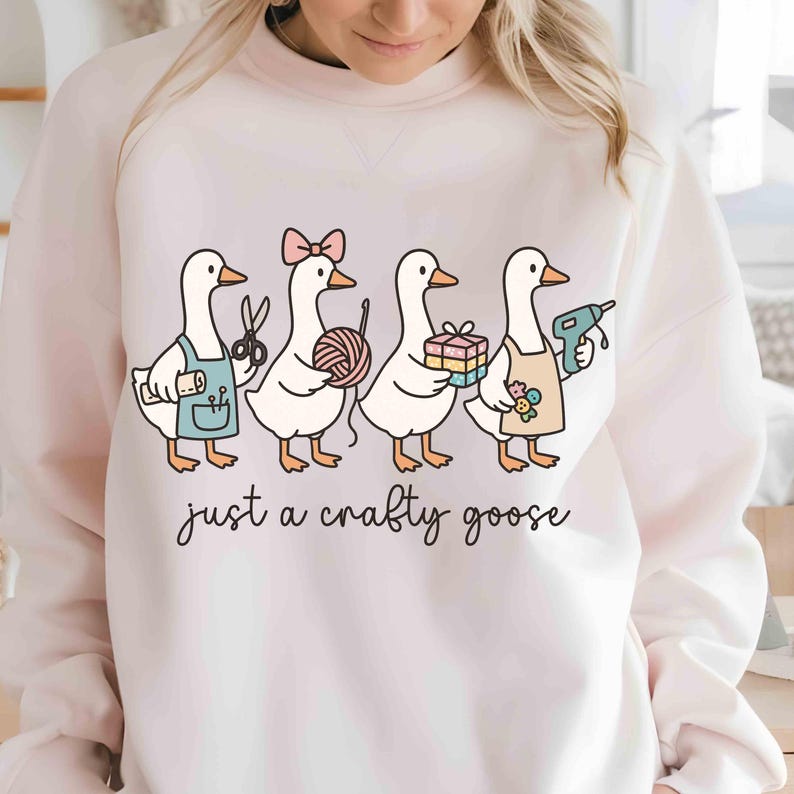 May include: A light pink sweatshirt featuring a whimsical illustration of four geese, each holding craft supplies. One goose has scissors, another yarn, a third a gift, and the last a glue gun. The text "just a crafty goose" is written below the geese.