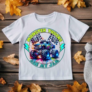 Puede incluir: Camiseta blanca con un gr&aacute;fico colorido de un cami&oacute;n monstruo. El dise&ntilde;o incluye banderas a cuadros y el texto "MONSTER TRUCKS ARE MY JAM". El cami&oacute;n es azul, verde y morado.