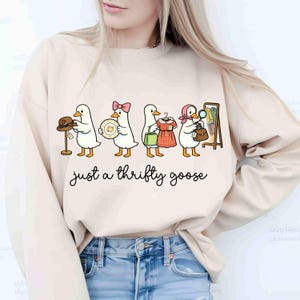 May include: Cream-coloured sweatshirt with a cartoon design of five geese, each carrying second-hand items such as a hat, plate, dress, and handbag. The text "just a thrifty goose" is written in a cursive font below the geese.