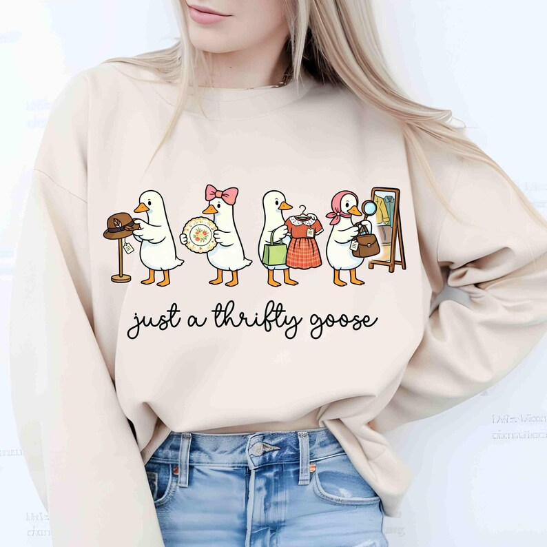 May include: Cream-coloured sweatshirt with a cartoon design of five geese, each carrying second-hand items such as a hat, plate, dress, and handbag. The text "just a thrifty goose" is written in a cursive font below the geese.